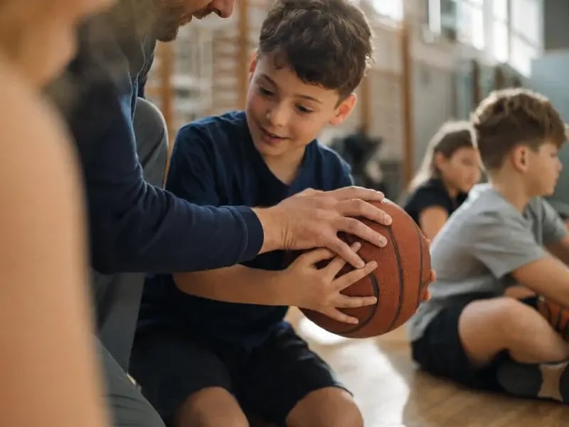 Trainer zeigt einem Grundschulkind beim Basketball-AG-Training den richtigen Griff am Ball, weitere Kinder sitzen im Hintergrund