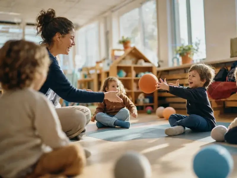 Erzieherin spielt mit Kita-Kindern im Sitzkreis bei Wolfis Ballschule und rollt einem lachenden Jungen einen weichen Ball zu